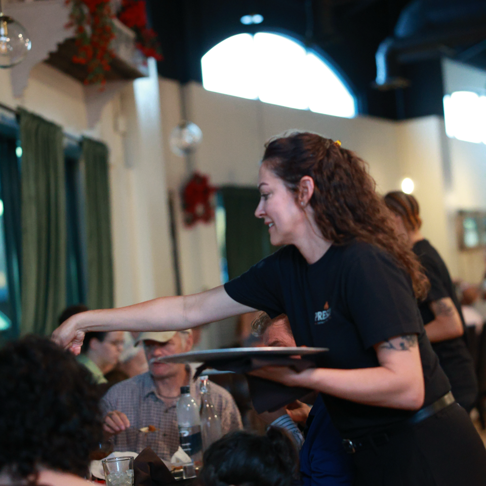 Server carries a tray through a busy, warmly decorated restaurant dining room.