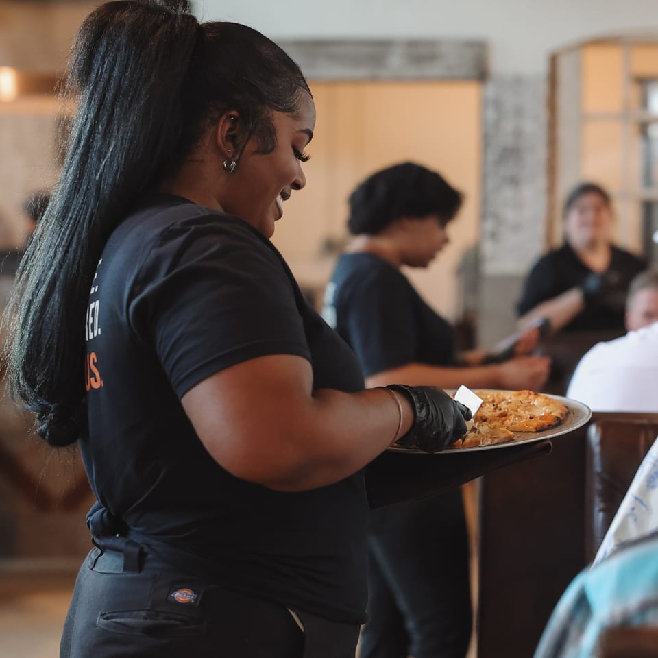 Restaurant server carries a tray of pizzas through a busy dining room while patrons sit at tables.