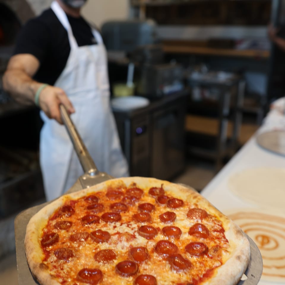 Pepperoni pizza on a metal serving tray, with a bearded restaurant worker holding a pizza peel in the background.