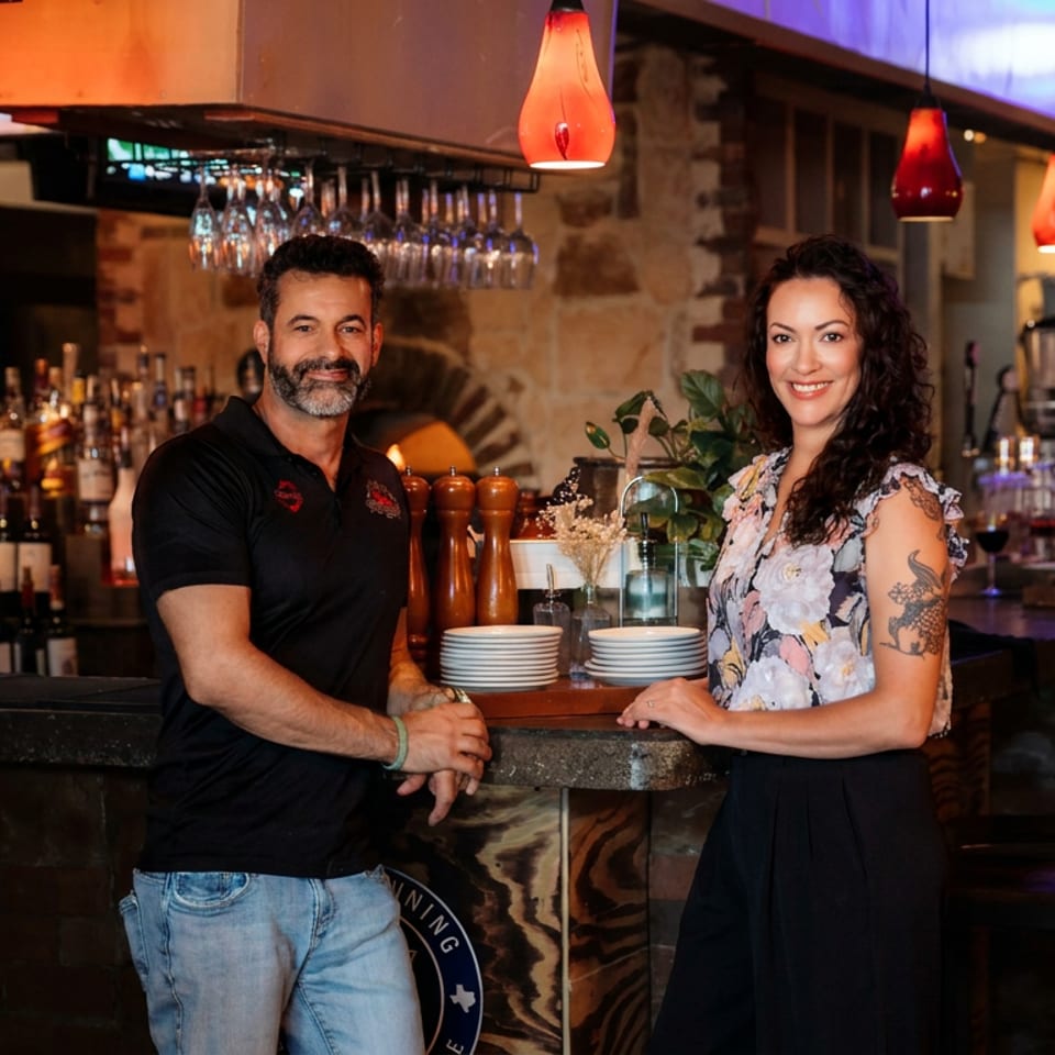 Two restaurant staff members pose at the bar amid hanging glassware, stacked plates, bottles, and warm pendant lighting.
