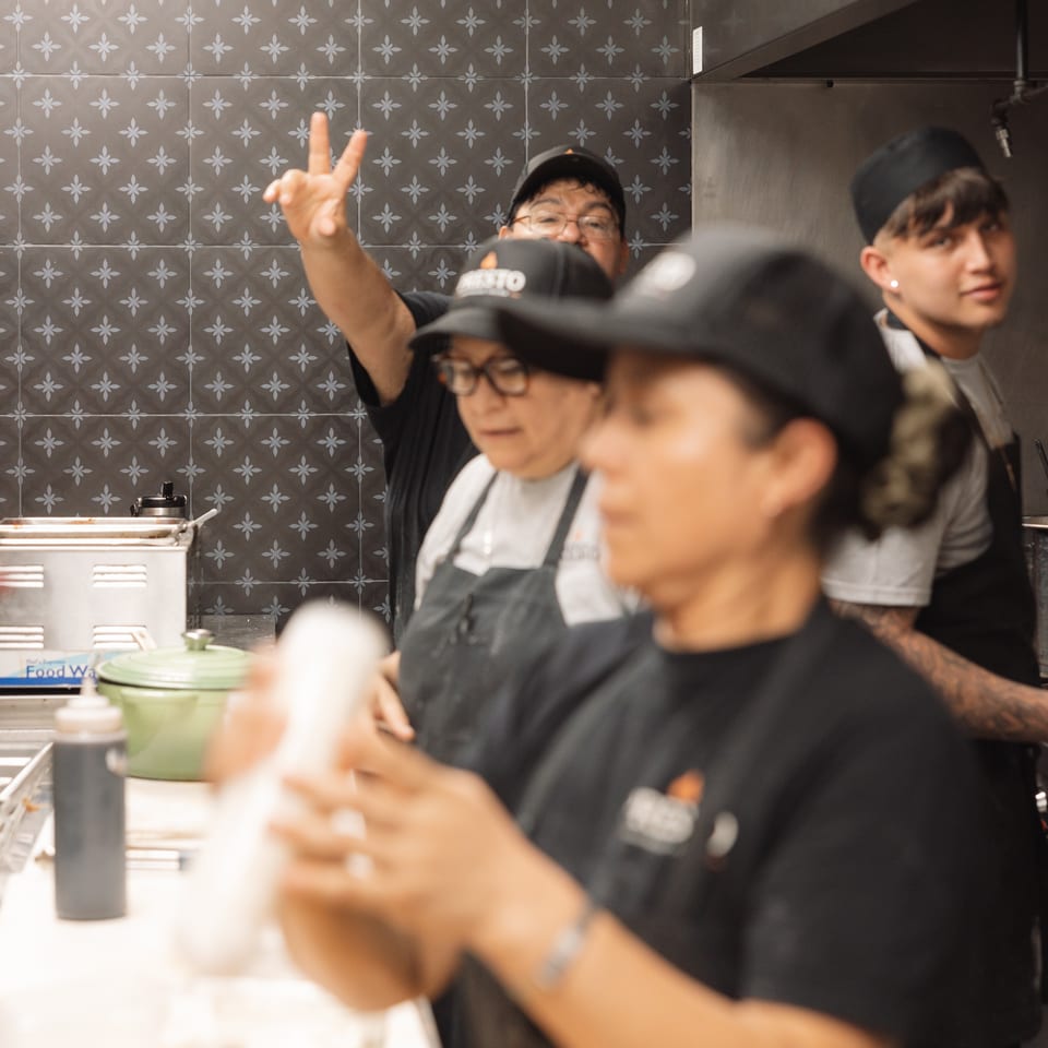 Restaurant staff gathered in a tiled kitchen, with one person flashing a peace sign beside food preparation counters.