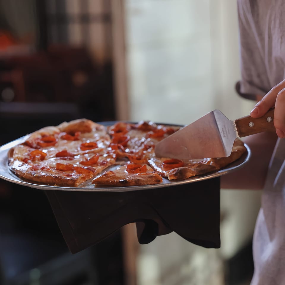 Server uses a pizza cutter to slice a pepperoni pizza on a metal serving tray.