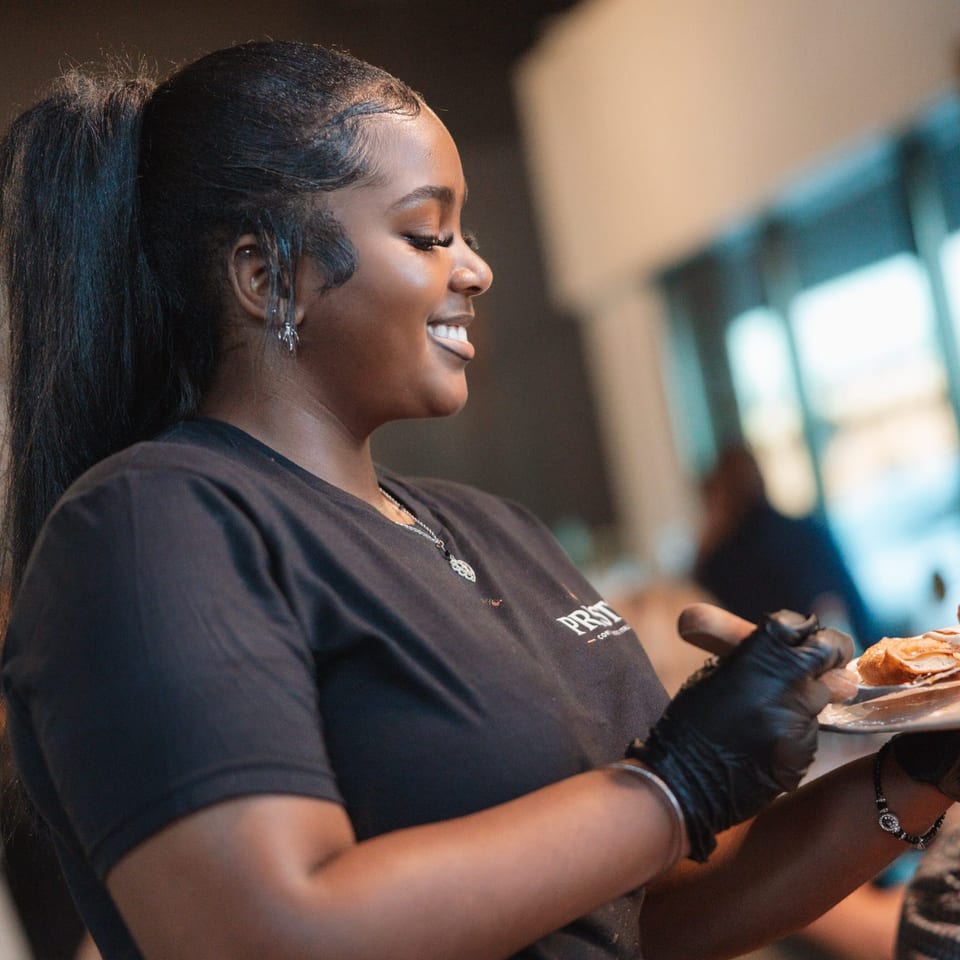 Smiling restaurant worker in black gloves presents a plated burger in a warmly lit dining room.