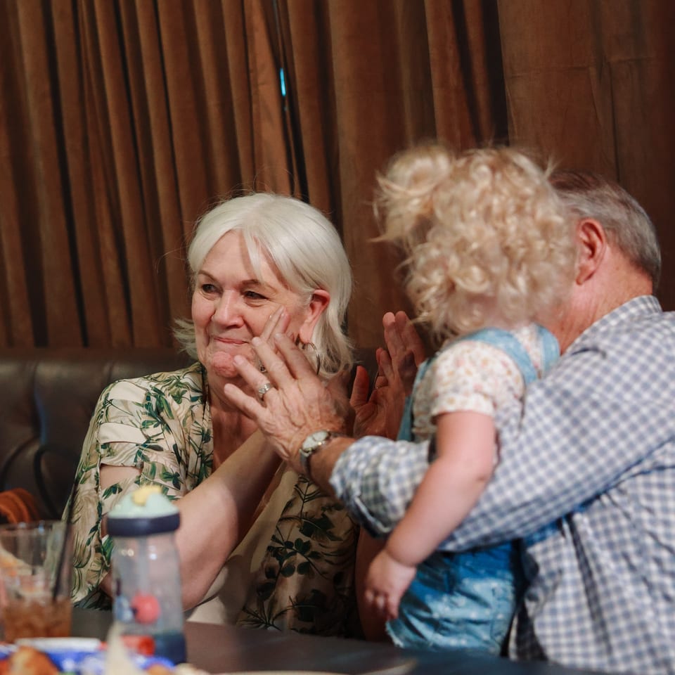Older adults applaud together at a restaurant table, with plates and drinks in the foreground.