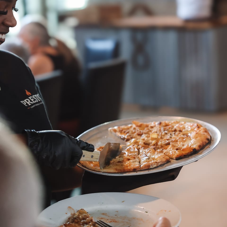 Restaurant server in gloves slices a cheesy baked dish onto a guest’s plate while diners sit nearby.
